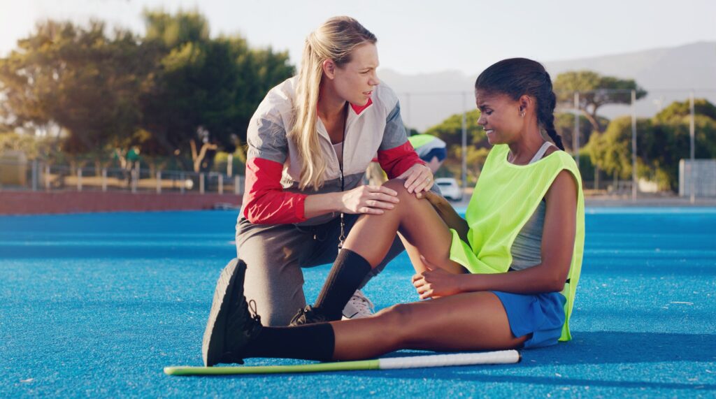 Women, hockey coach and knee injury girl on field together with support, solidarity or care at spor